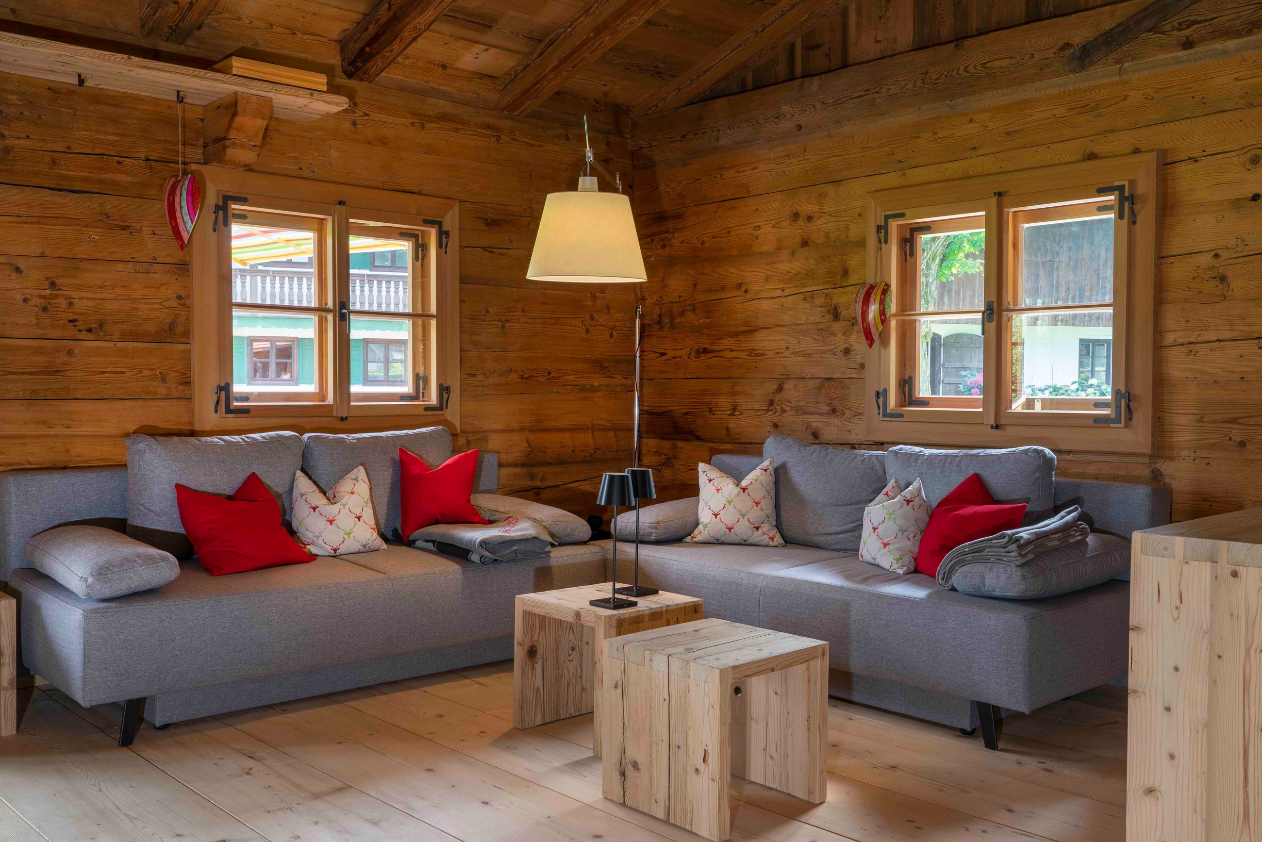 Gemütliches Wohnzimmer in einer Holzhütte mit zwei grauen Sofas, die mit roten und gemusterten Kissen verziert sind, Holztischen und einer Stehlampe.