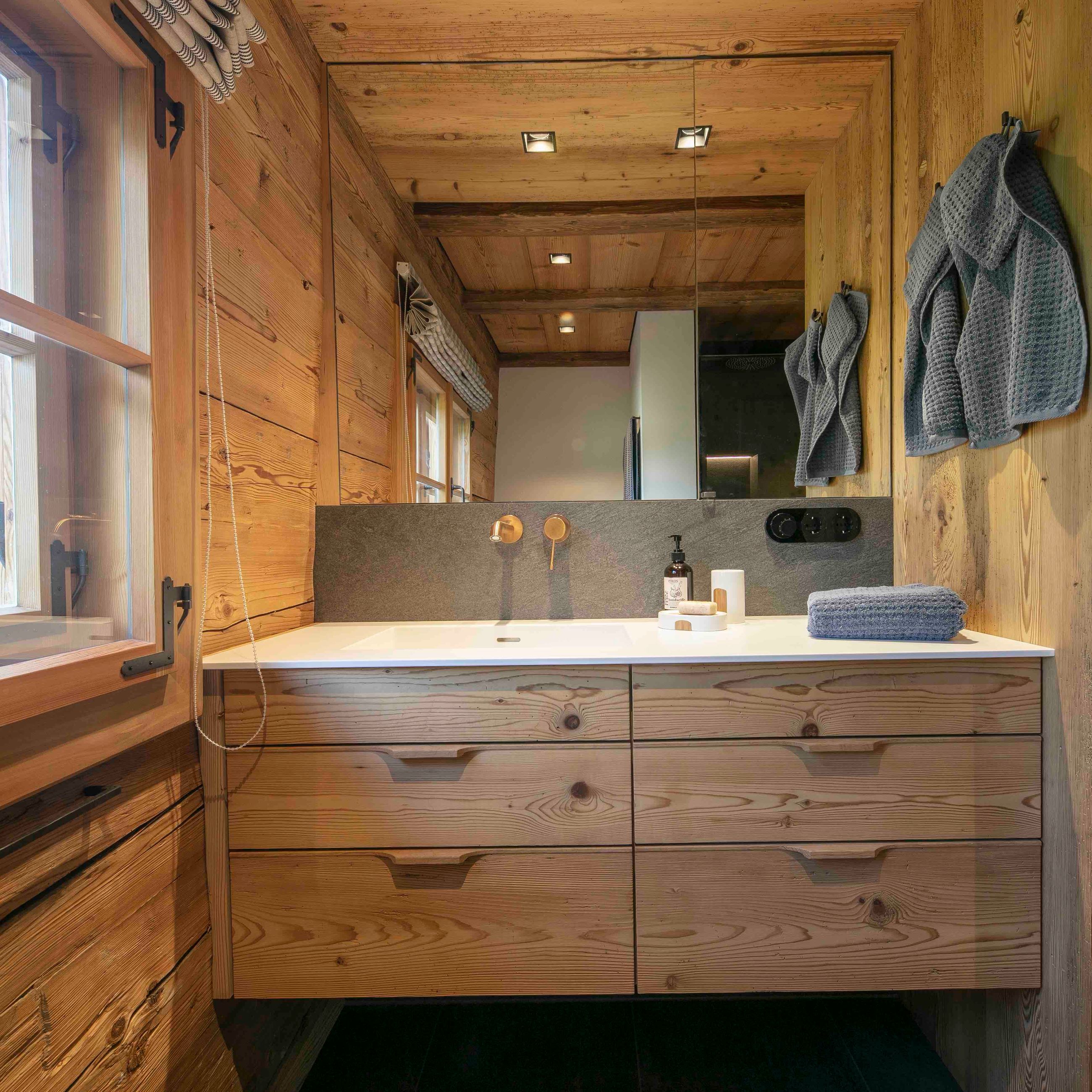 Rustikaler Waschtisch aus Holz mit weißer Arbeitsplatte, wandmontiertem Messingwasserhahn, grauen Handtüchern und einem großen Spiegel unter der Holzdecke.