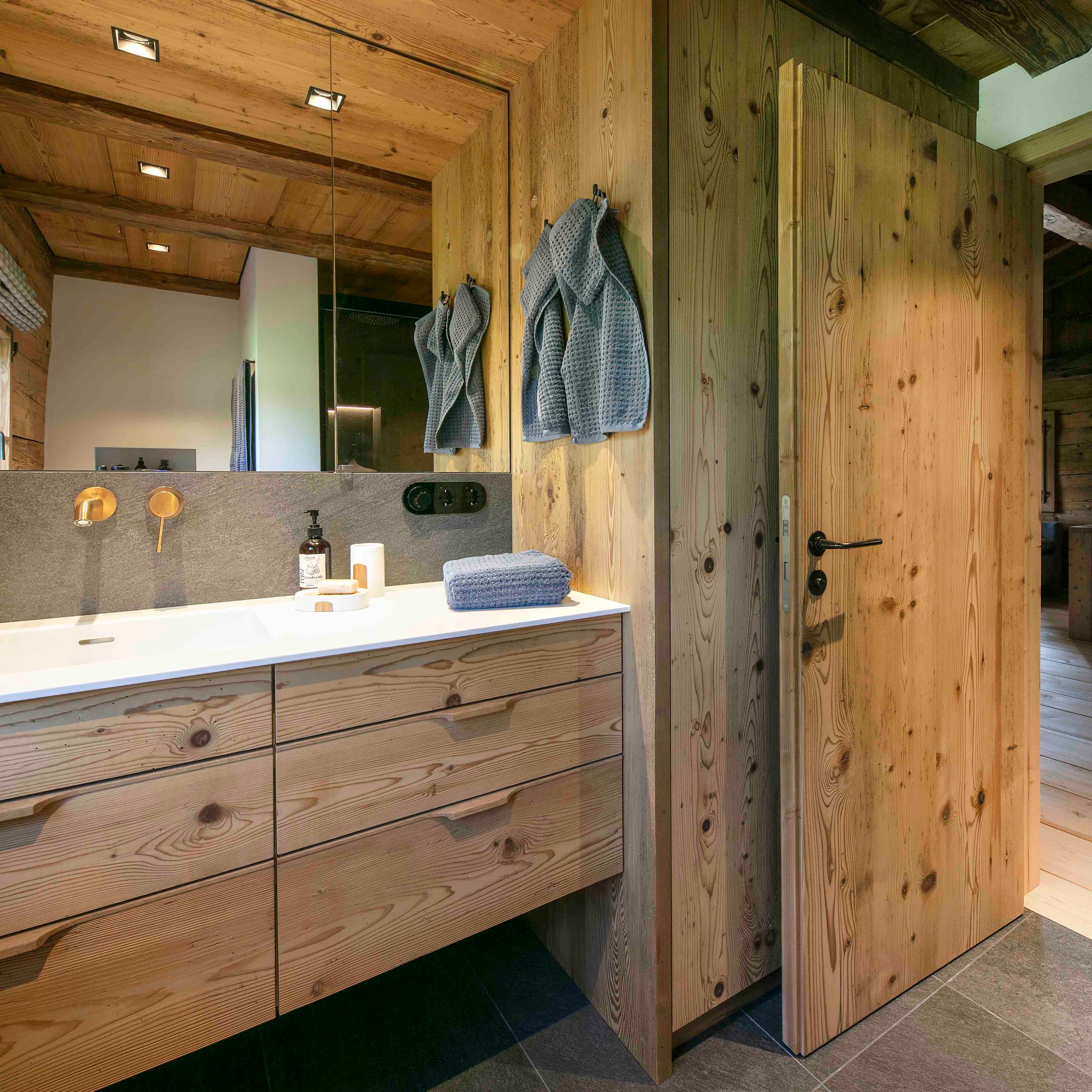 Badezimmer mit Holzschränken, einem weißen Waschbecken, grauen Handtüchern an der Wand und einer leicht geöffneten Holztür.