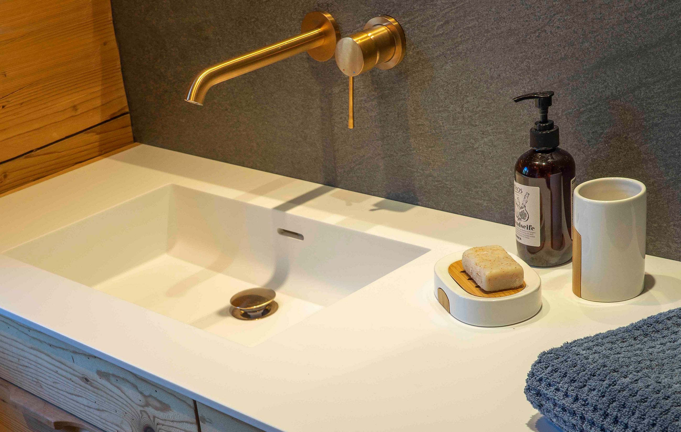 Modernes Waschbecken mit goldenem Wandwasserhahn, Seifenstück auf einer Seifenschale, Flüssigseifenspender, Zahnbürstenhalter und einem gefalteten blauen Handtuch.