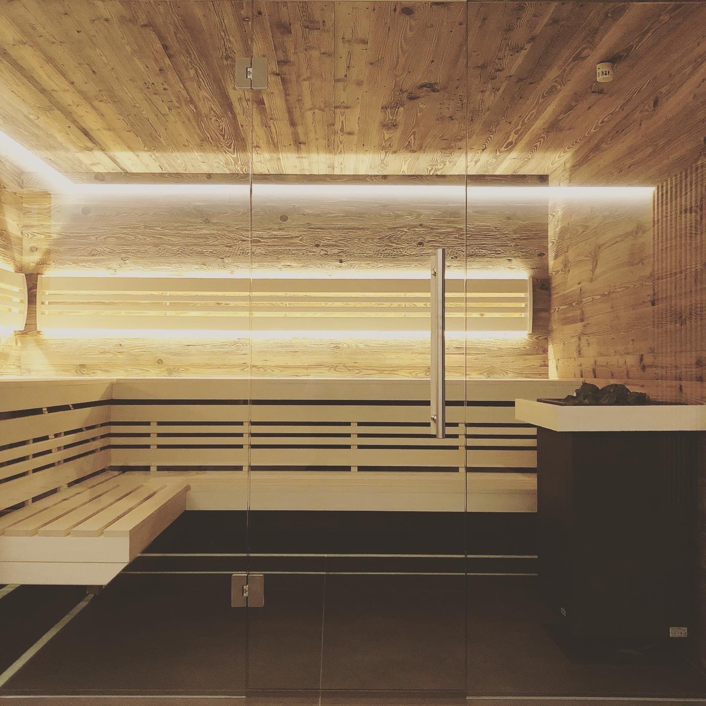 Modernes Sauna-Interieur mit Holzwänden und -decke, weißen Lattenbänken, indirekter warmer Beleuchtung und einem Ofen mit Saunasteinen.