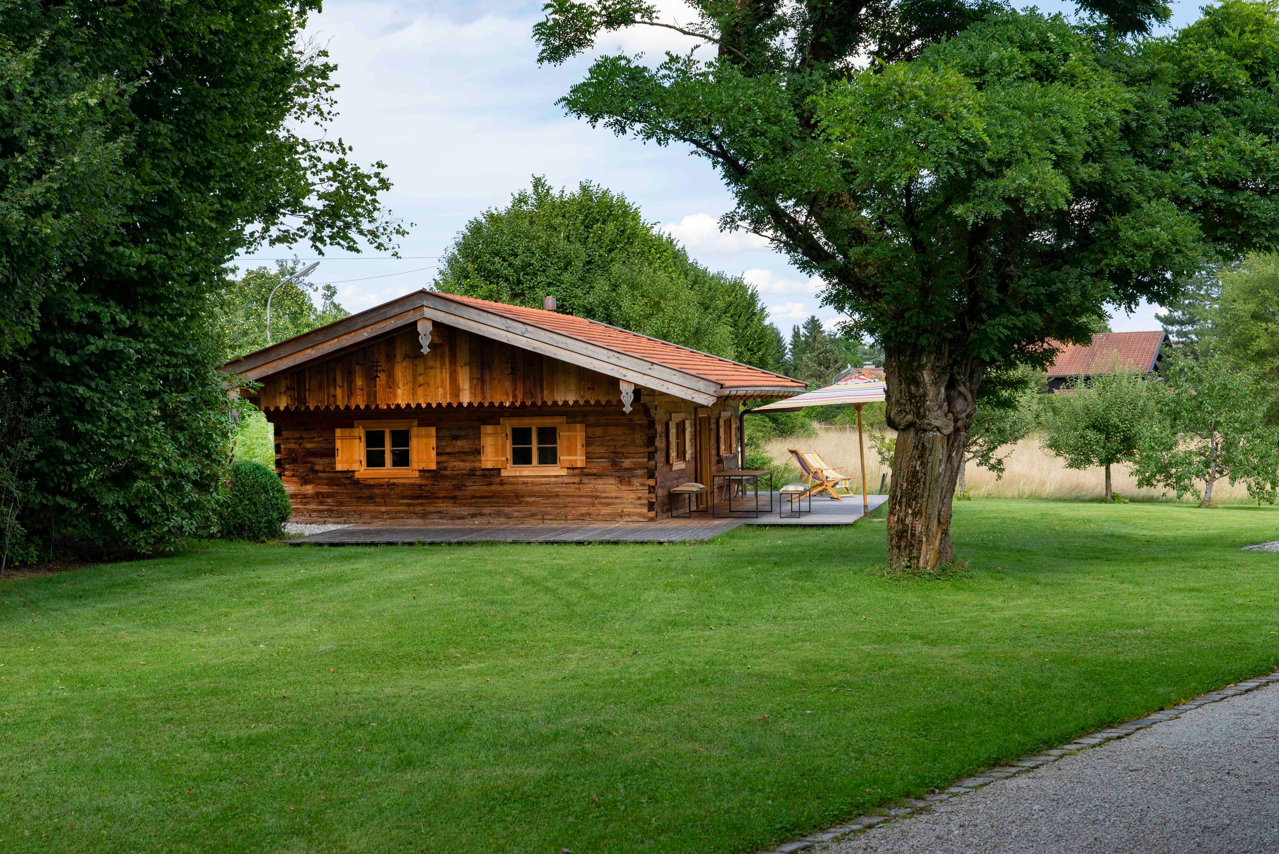 Holzhütte mit Fensterläden und Gartenmöbeln unter einem Sonnenschirm in einem grünen Garten mit Bäumen.