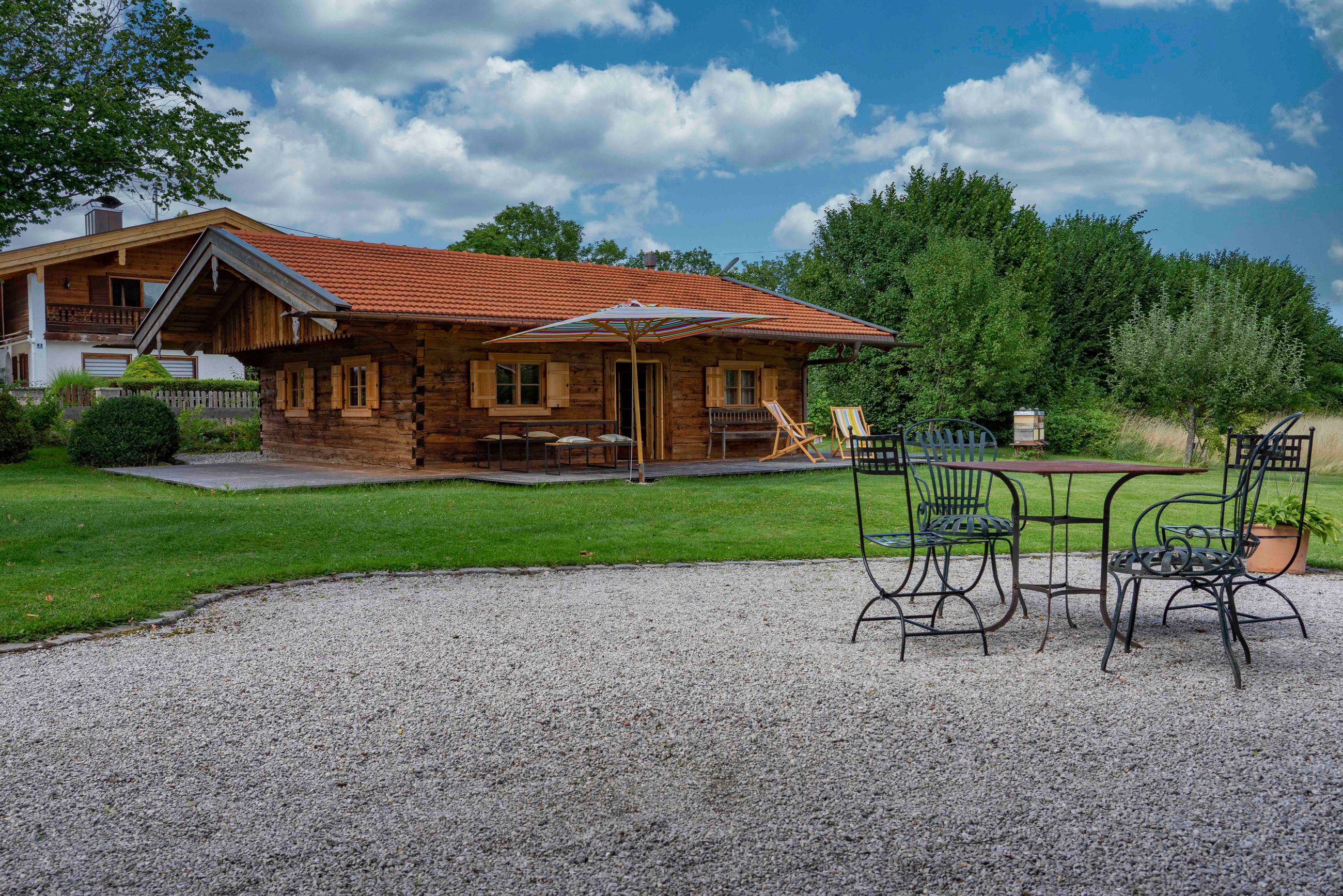 Außenansicht einer Holzhütte mit rotem Ziegeldach, einer Terrasse mit Liegestühlen und einem großen Sonnenschirm sowie einem Metalltisch mit vier Stühlen auf einem Kiesbereich.