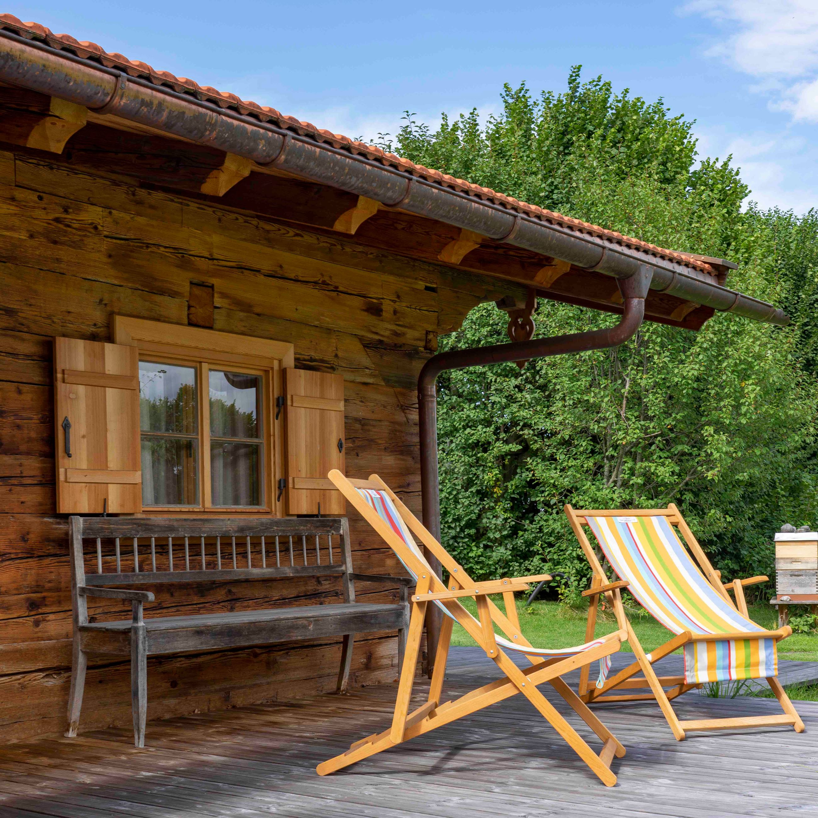 Holzterrasse mit zwei gestreiften Liegestühlen und einer rustikalen Holzbank vor einer Blockhütte mit Fensterläden.