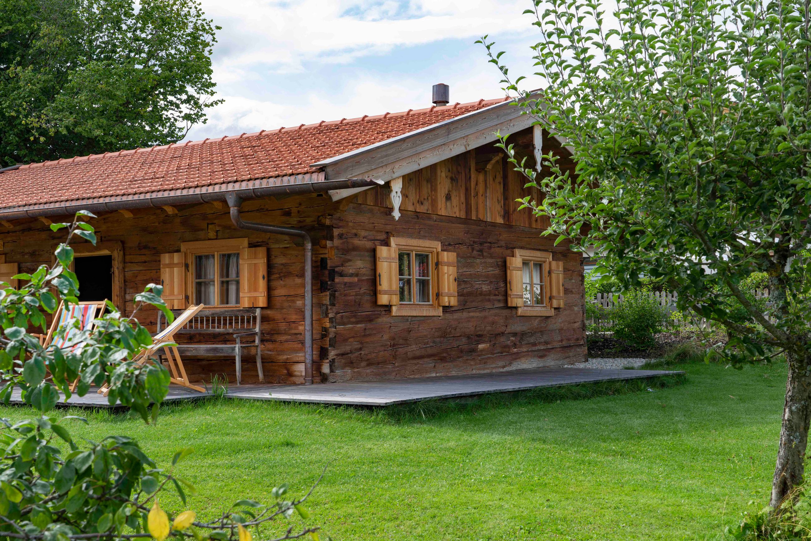Rustikale Holzhütte mit rotem Ziegeldach, drei Fensterläden, einer Bank und zwei gestreiften Liegestühlen auf einer Holzveranda, umgeben von grünem Gras und Bäumen.