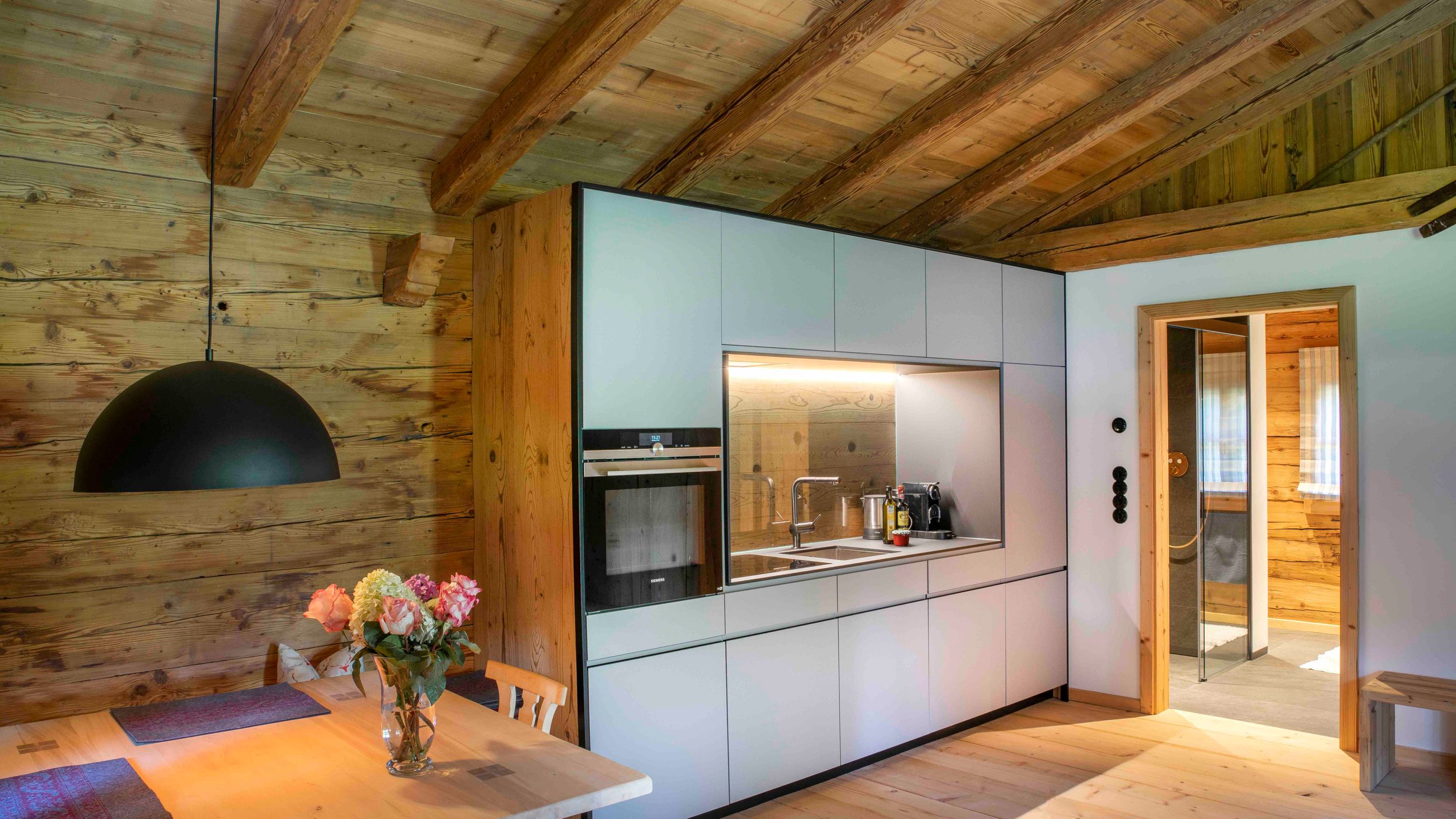 Gemütliche, moderne Küche mit weißen Schränken, Einbauofen, Holzdecke und -wänden, Holztisch mit Blumenvase und schwarzer Pendelleuchte.