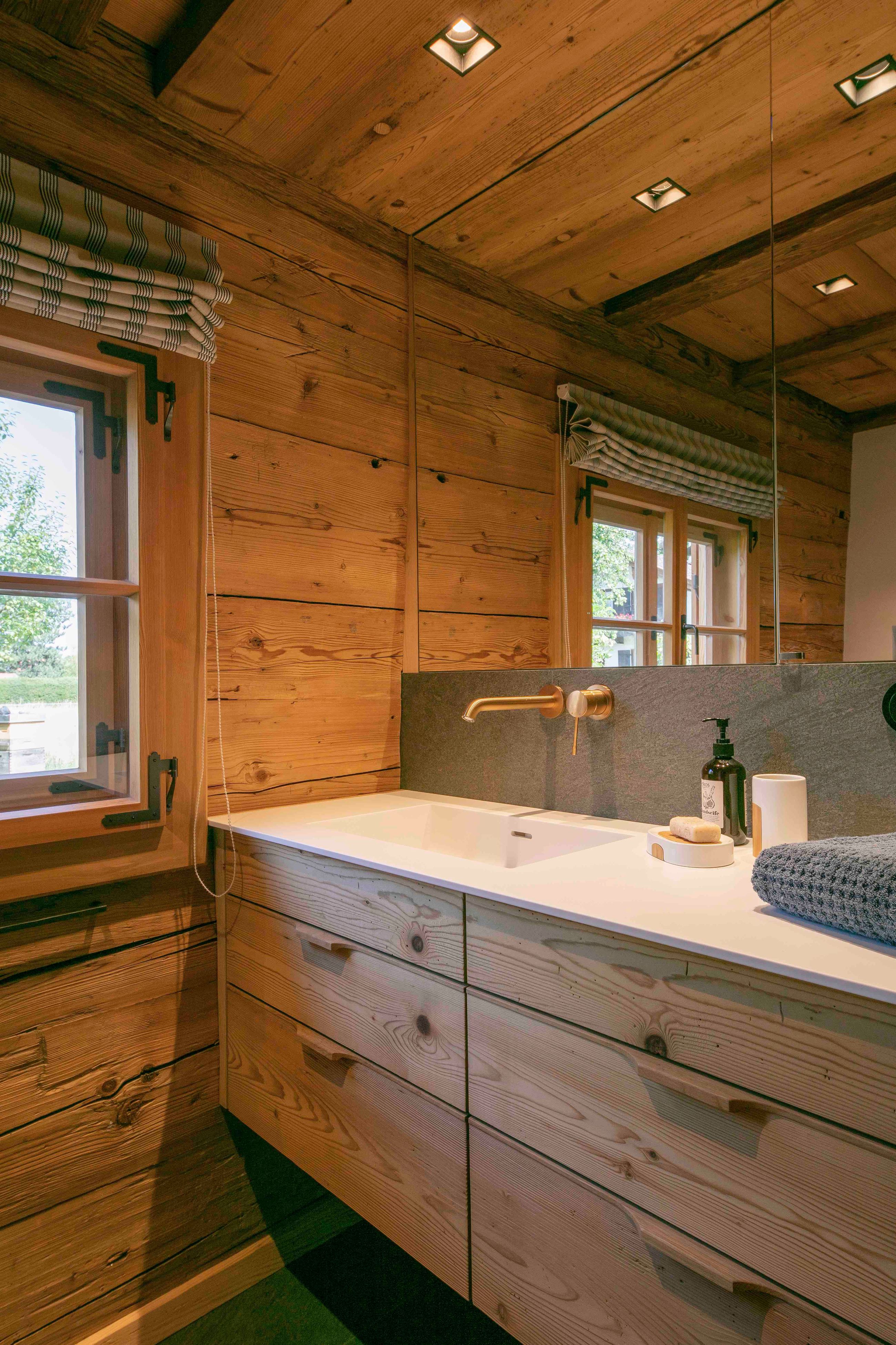 Rustikaler Waschtisch aus Holz mit weißer Arbeitsplatte, Messingarmatur und ordentlich angeordneten Seifen und Handtüchern unter einem Fenster mit gestreiften Raffrollos