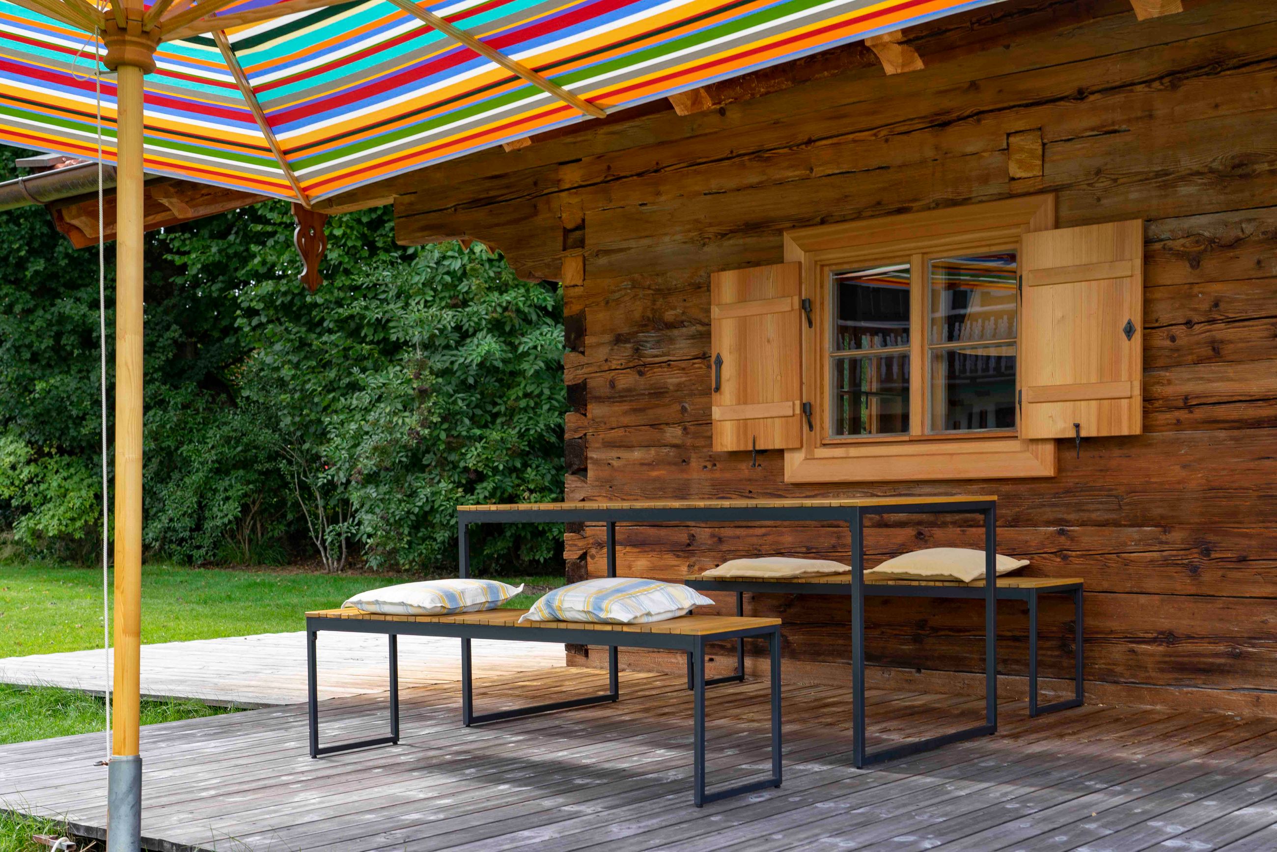 Holzterrasse im Freien mit modernen Bänken und Tisch mit Metallgestell, bunt gestreiftem Sonnenschirm und einer rustikalen Holzhütte mit offenen Fensterläden.